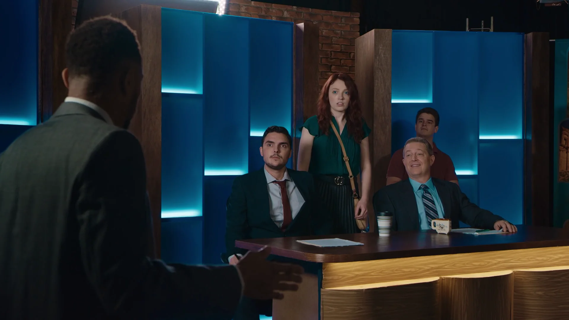 The Bad News ensemble gathers around a news desk on a blue-lit set.