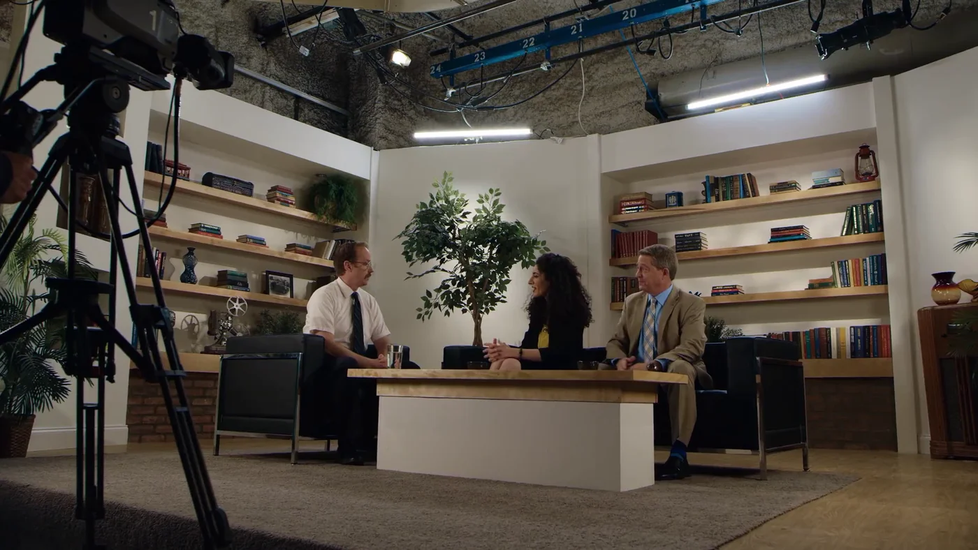 A wide shot of a television interview set with cameras and overhead lighting in Bad News.