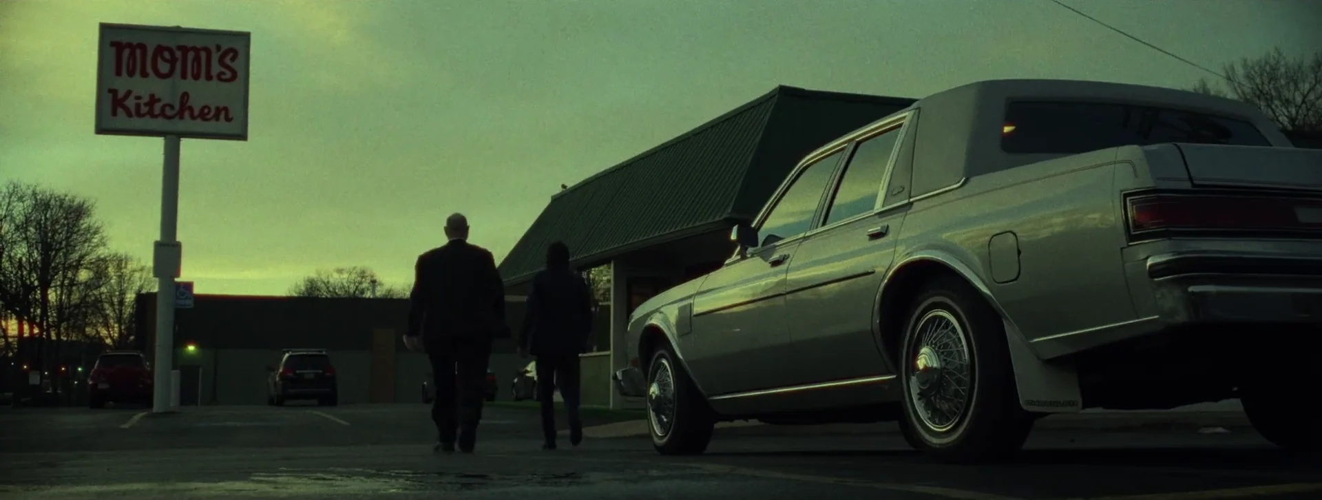 Two figures in dark coats walk toward a roadside diner called Mom’s Kitchen at dusk in Justice for Peanut.