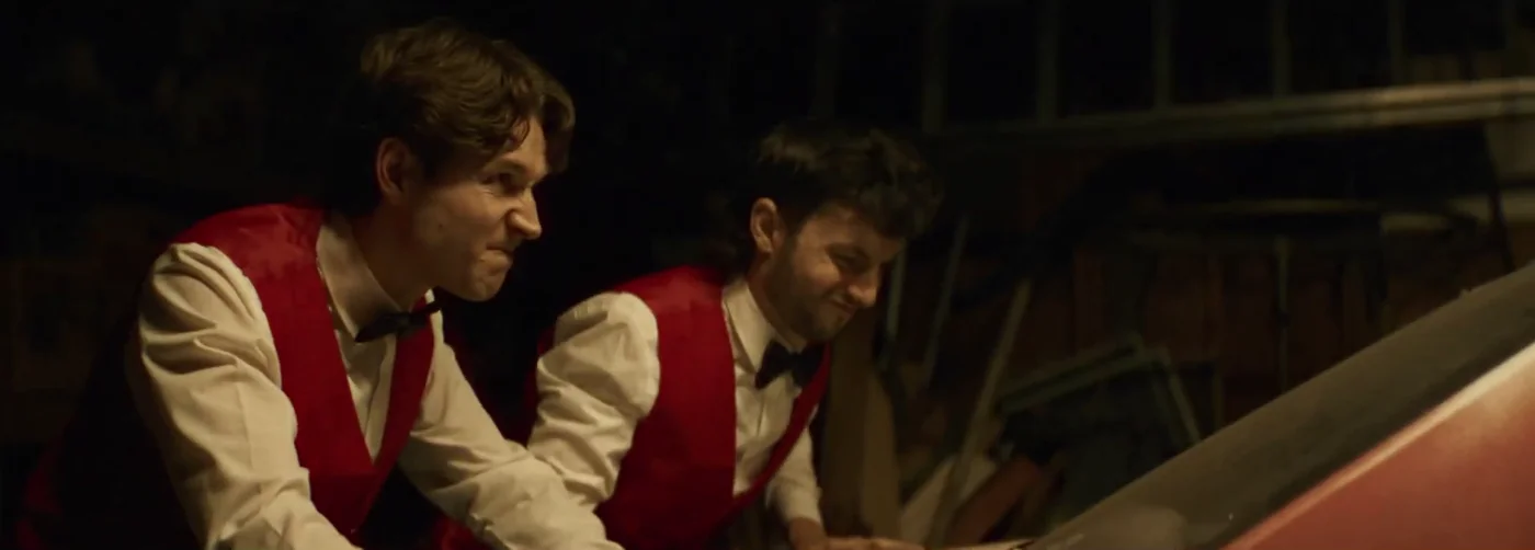 Two men in red vests and bow ties strain against a red car in a dark garage in Kings of KC.