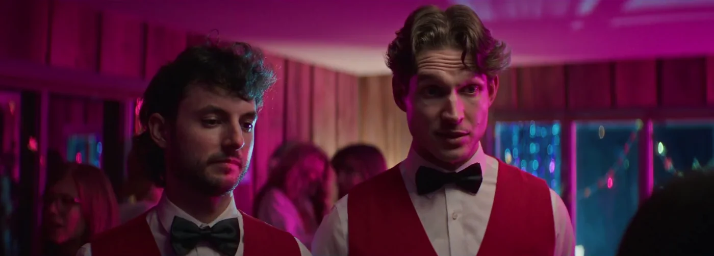 Two men in red vests and bow ties at a pink-lit party in Kings of KC.