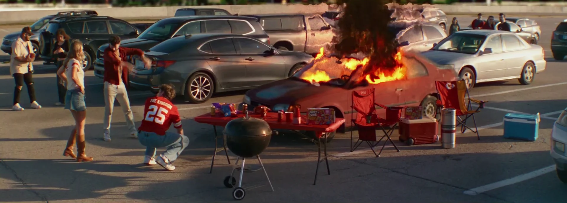 A car burns in a stadium parking lot as tailgaters in red keep the party going in Kings of KC.