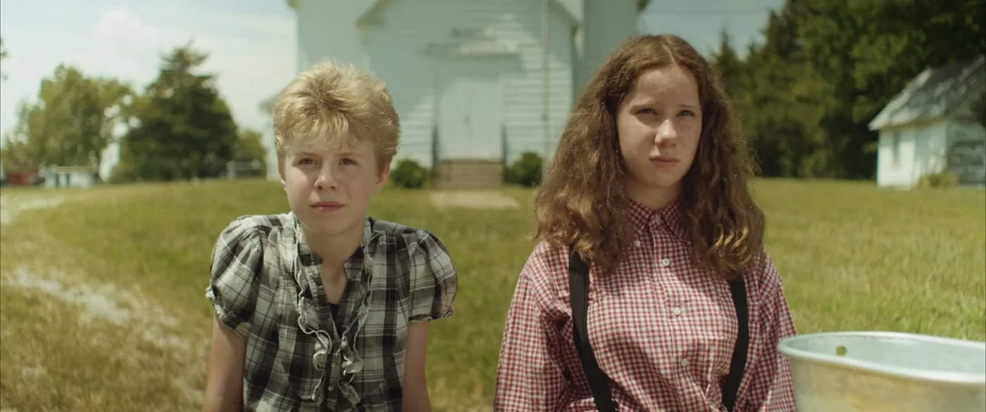 The two children sit in a sunny field in front of a white clapboard building in The Escape of Tina Kleinfelder.