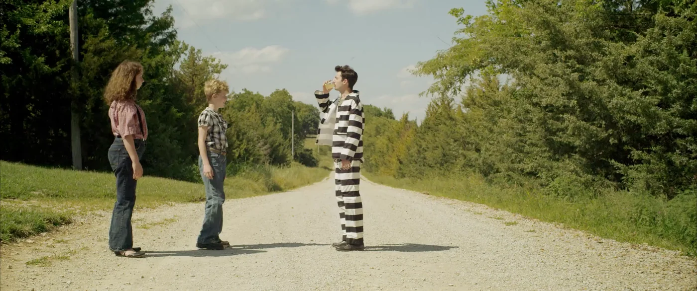 The man in stripes drinks lemonade on a gravel road as the two children watch in The Escape of Tina Kleinfelder.