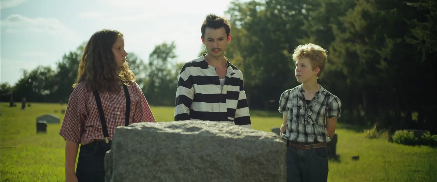 The man in stripes and the two children stand at a gravestone in a grassy cemetery in The Escape of Tina Kleinfelder.